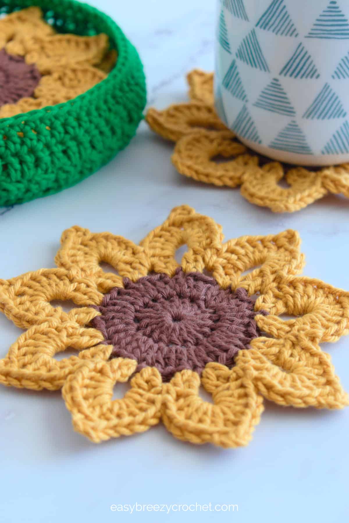Close up of a crochet sunflower coaster with a cup on a coaster in the background.