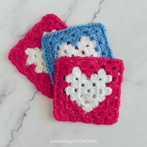 Three crocheted heart granny squares, two are dark pink and one blue.