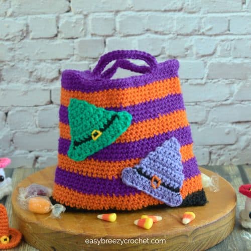 A orange an purple Halloween trick or treat bag on a round board with candy scattered around.
