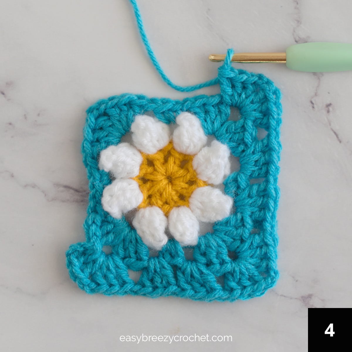 A partially compete blue crochet granny square with a white flower.