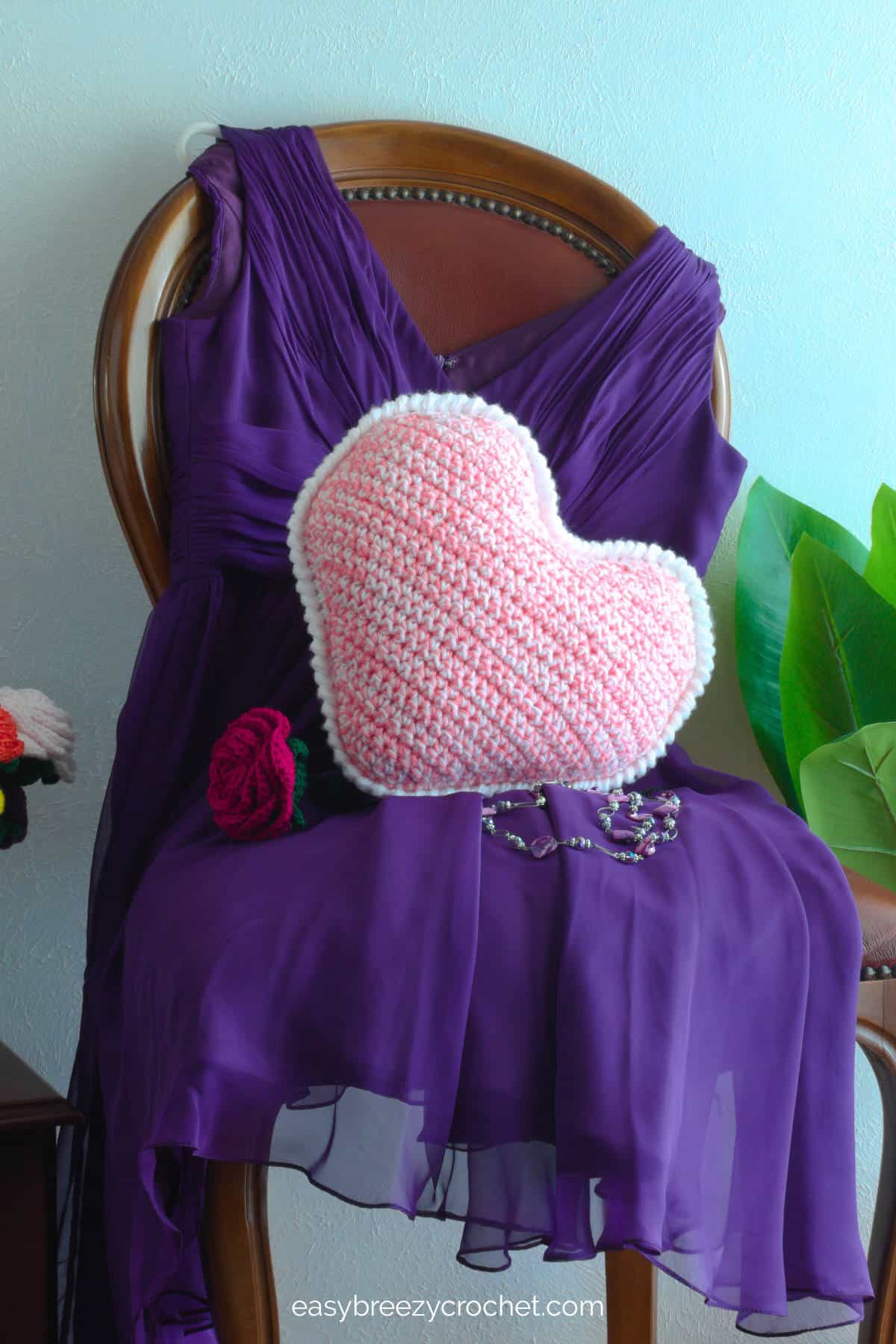 A pink and white crochet heart pillow on a purple dress on a chair.