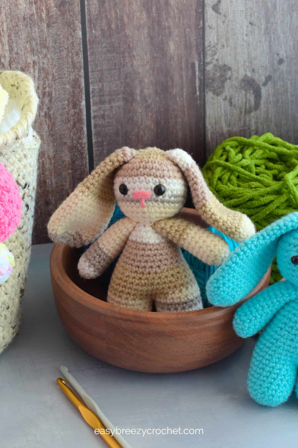 A brown multi colored bunny in a wooden bowl with crochet hooks in the foreground.